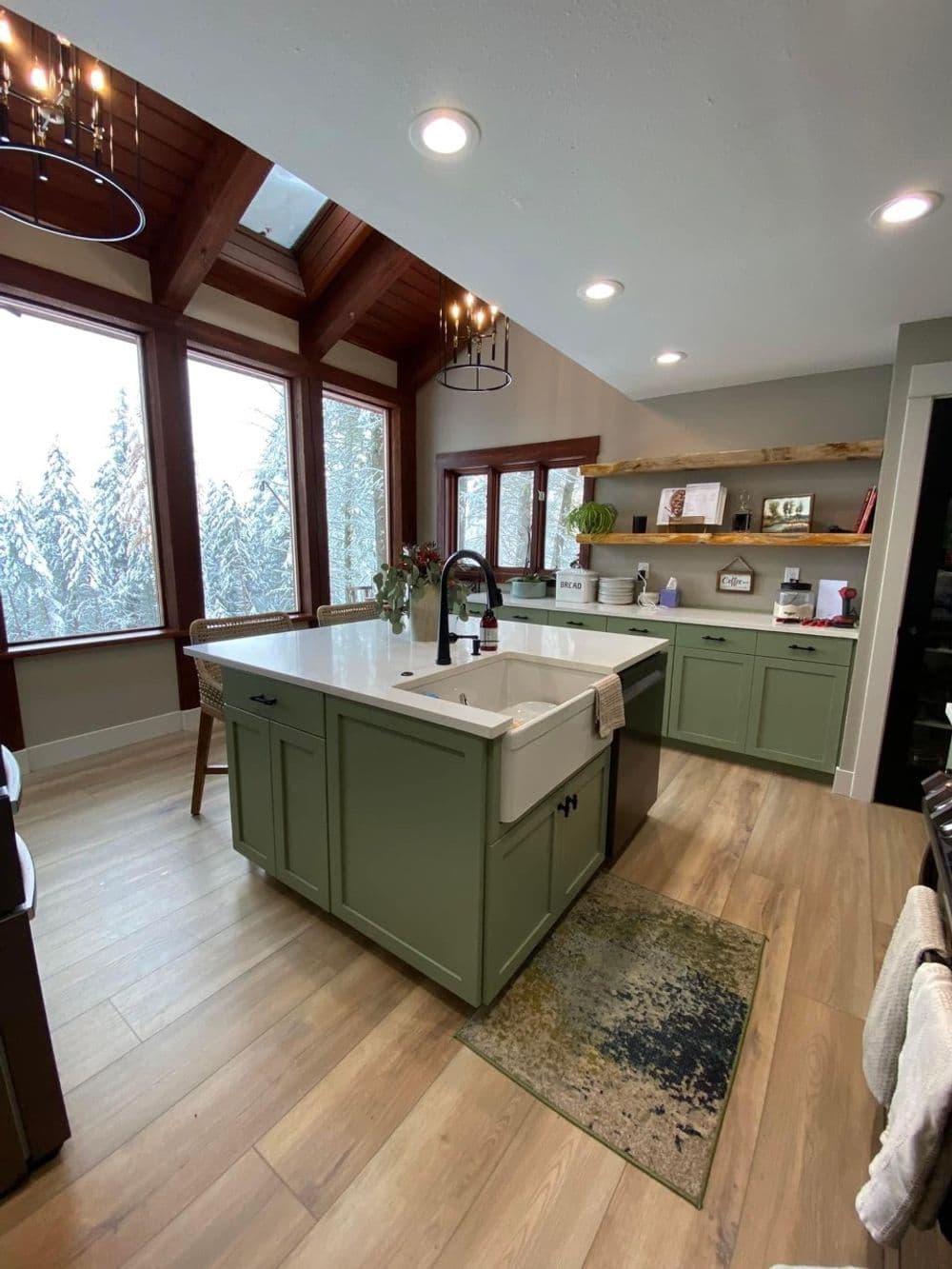 Modern kitchen with green cabinets, white farmhouse sink, large windows, and wooden accents.